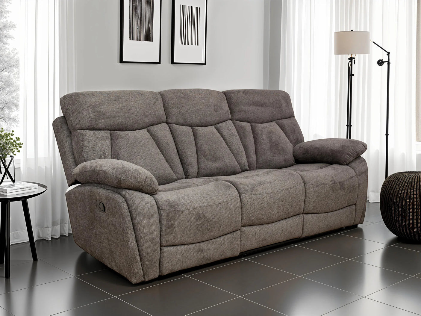 A three-seat gray reclining sofa with plush padded cushions, shown in a modern living room setting with dark tiled flooring, white curtains, and a floor lamp.