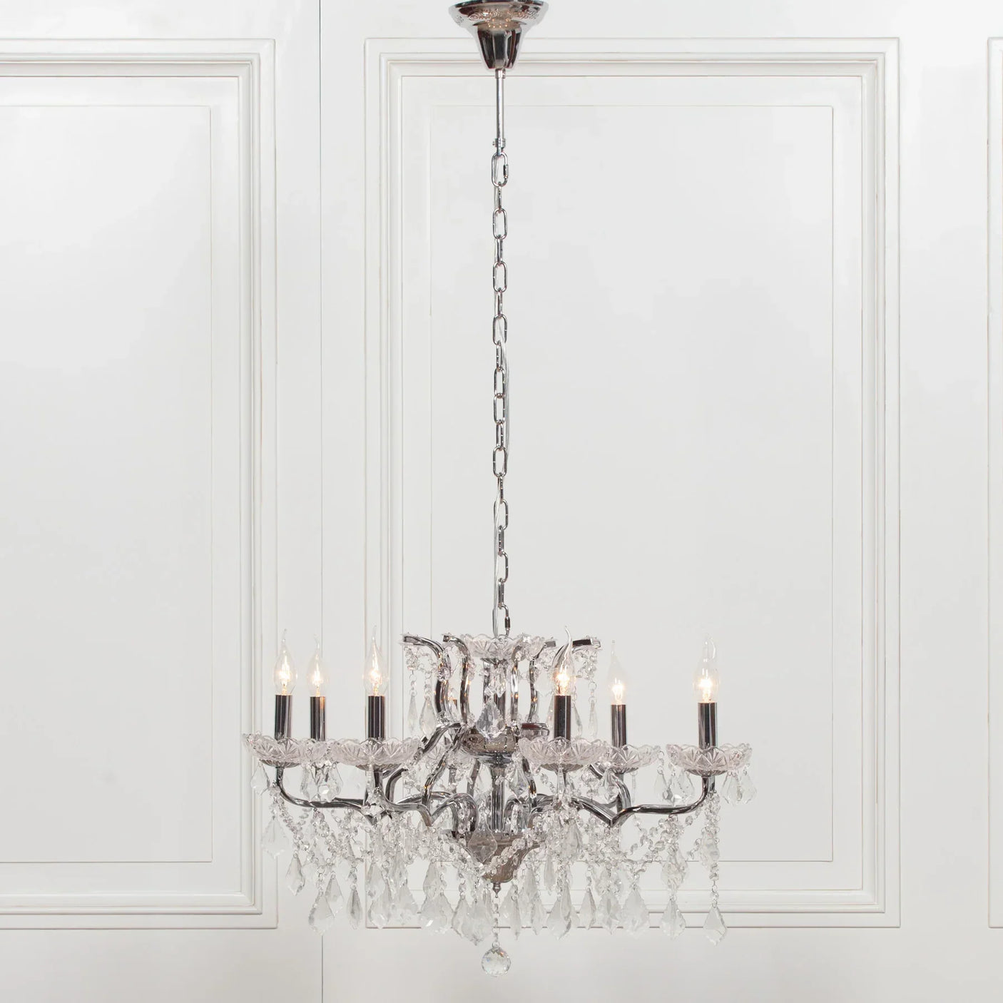 Crystal chandelier with chrome frame and candle-style bulbs hanging in front of white panel wall
