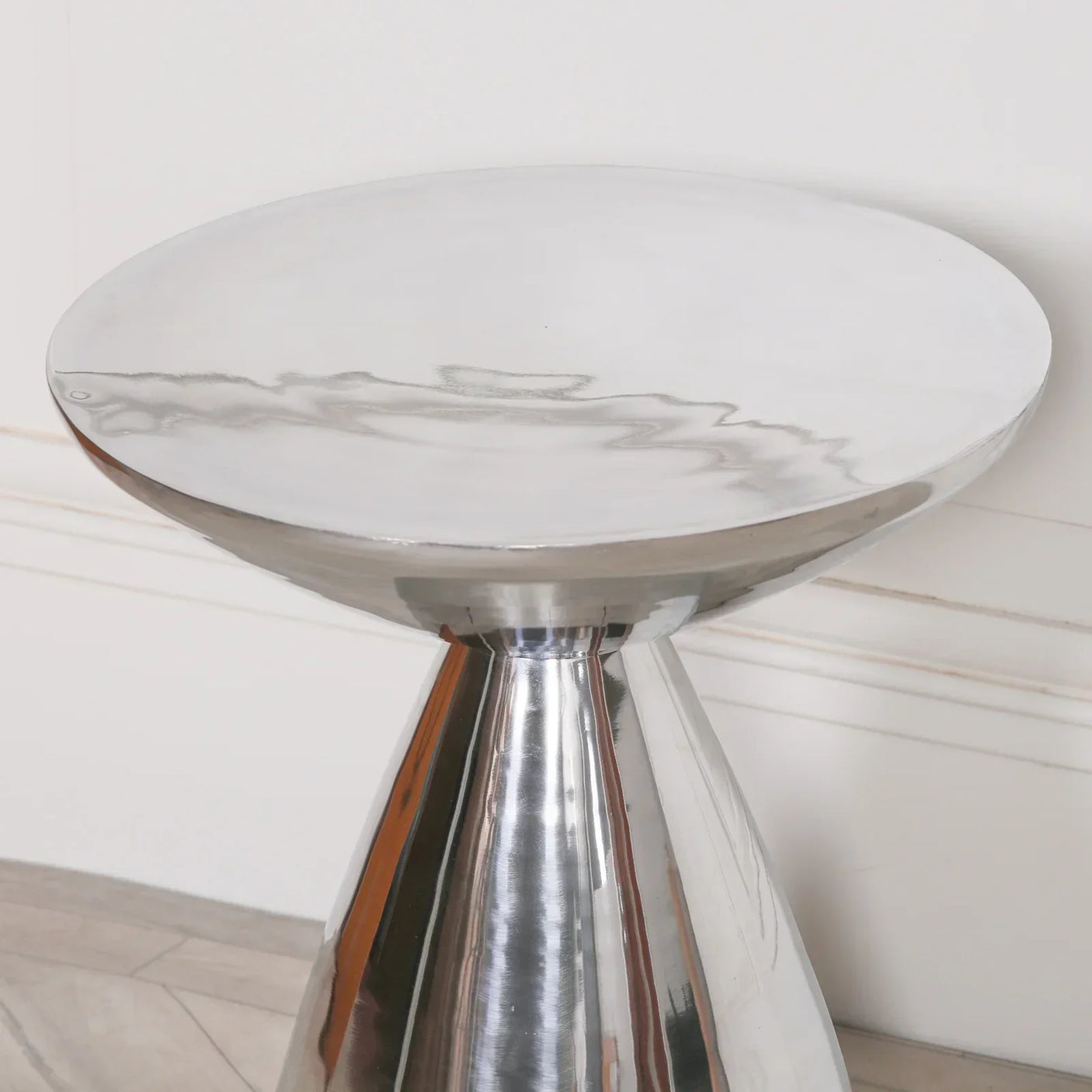 Modern round metal side table with polished silver finish, angled against a white wall