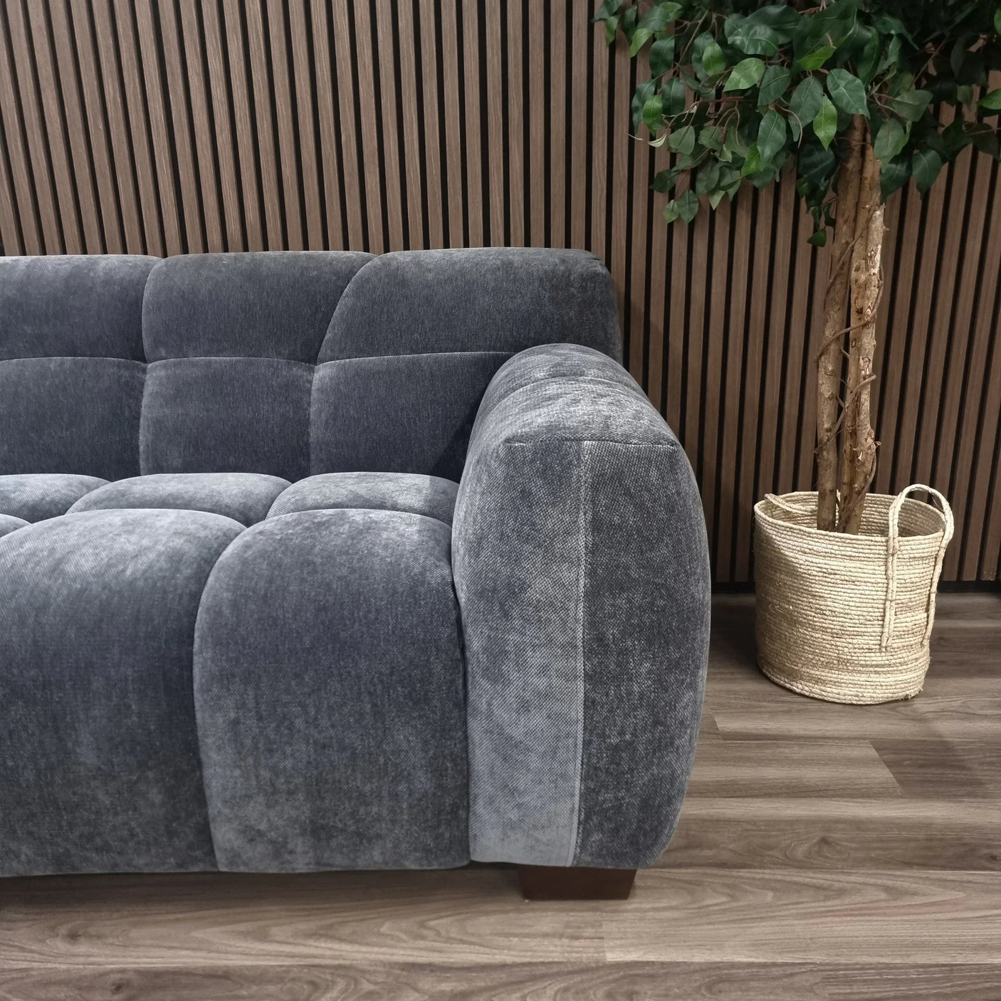 Grey fabric corner sofa with tufted cushions beside potted plant, wood panel backdrop