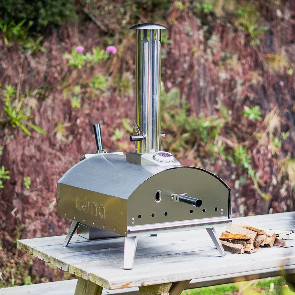 Stainless steel outdoor pizza oven with chimney on wooden table, firewood stacked beside