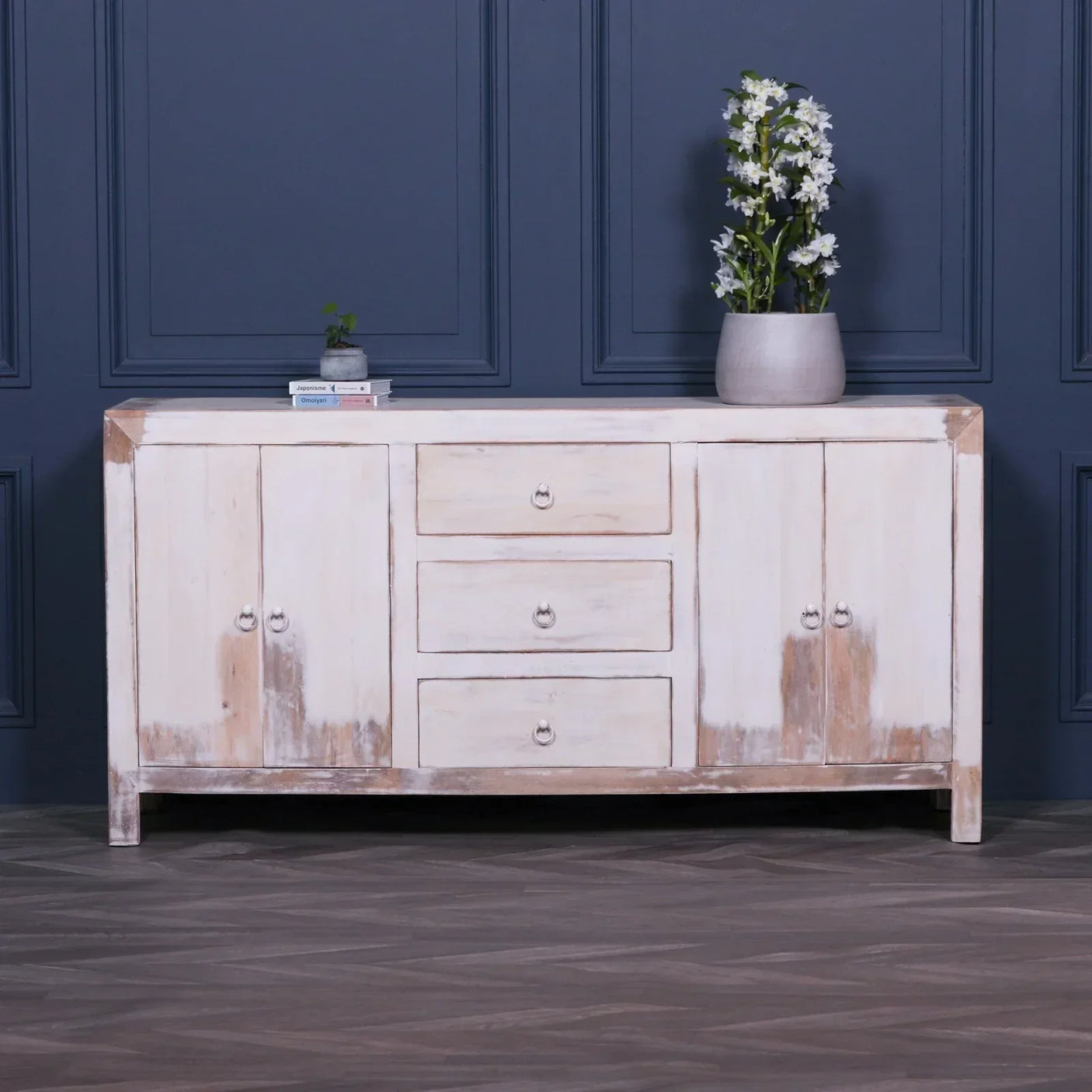 Distressed white wooden sideboard with three drawers and cupboards against blue wall