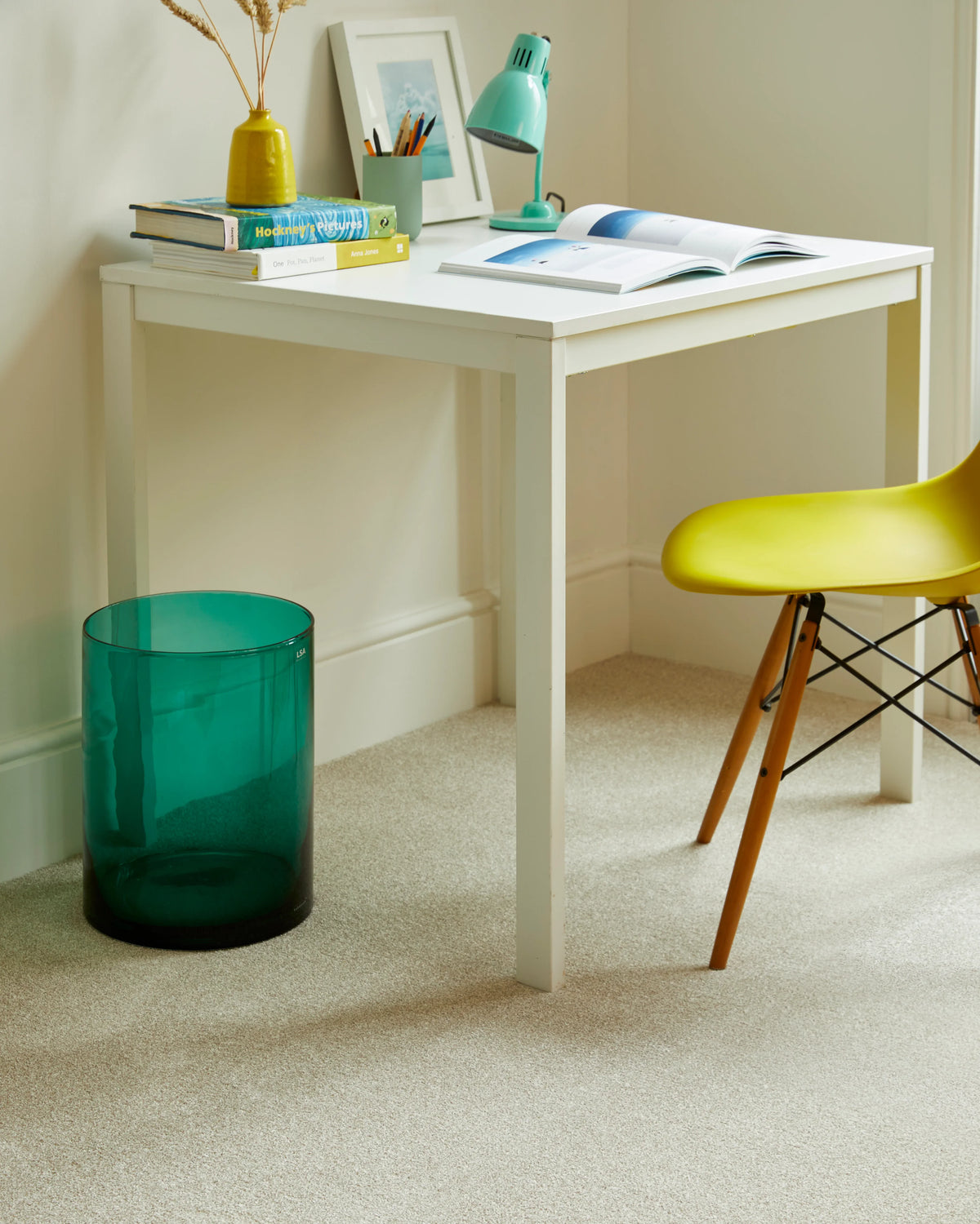 White desk with open book and green lamp in a cozy study corner with beige carpet.