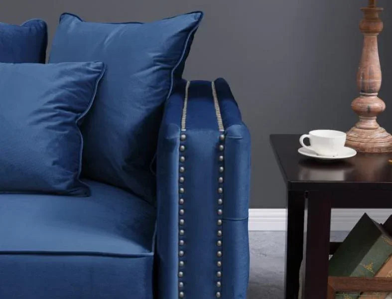 Blue velvet sofa with nailhead trim beside dark wood side table and stacked books