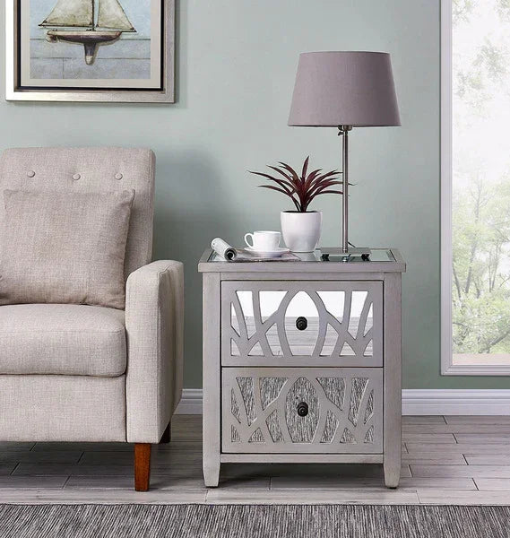 Grey mirrored bedside table with two drawers beside beige fabric armchair, lamp on top
