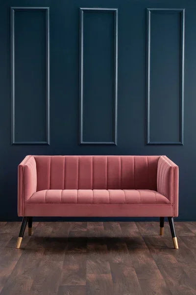 Pink velvet sofa with gold-tipped legs on dark wood floor against navy panel wall