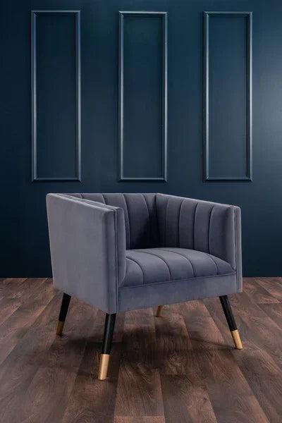 Grey velvet accent chair with gold-tipped legs on wood floor against dark panel wall