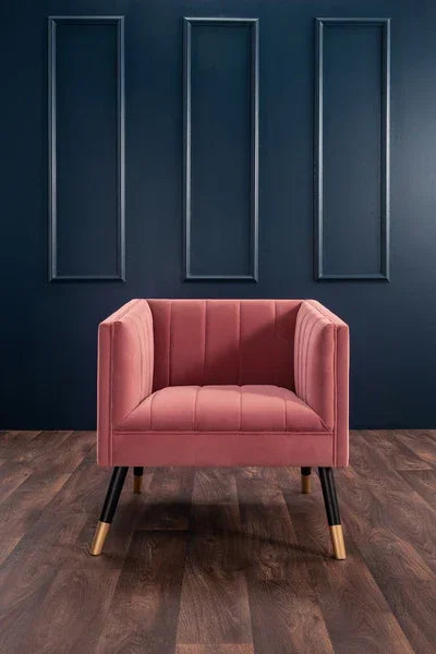 Blush pink velvet accent chair with gold metal legs on wood floor against dark wall
