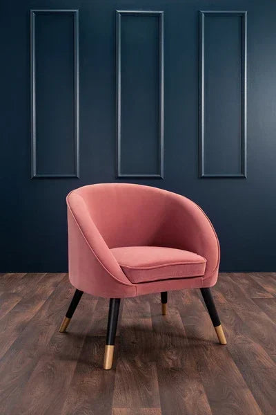 Pink velvet accent chair with gold-tipped legs on wooden floor against dark paneled wall