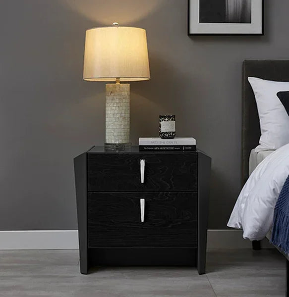 Black wood bedside table with two drawers, lamp, and books next to bed