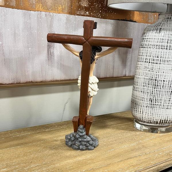 Crucifix figurine on a wooden cross with a stone-effect base, angled view beside a table lamp