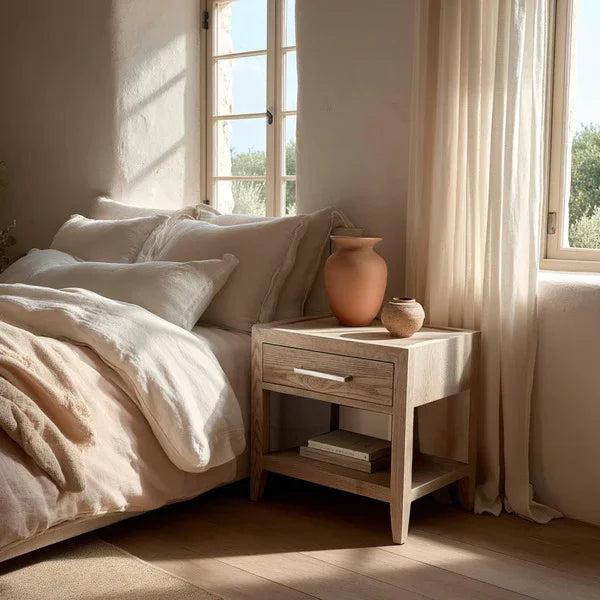 Light wood bedside table with drawer and shelf beside bed in sunlit bedroom