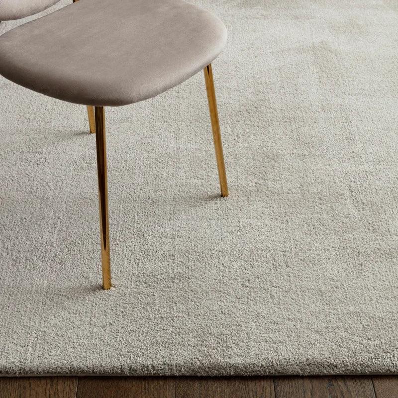 Light beige rug on wood floor with part of a velvet dining chair and gold metal legs visible