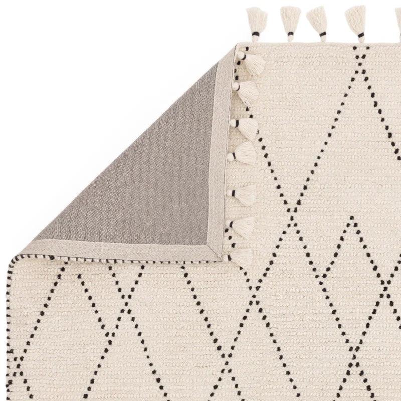 Cream woven rug with black diamond pattern and tassels, corner folded to show underside