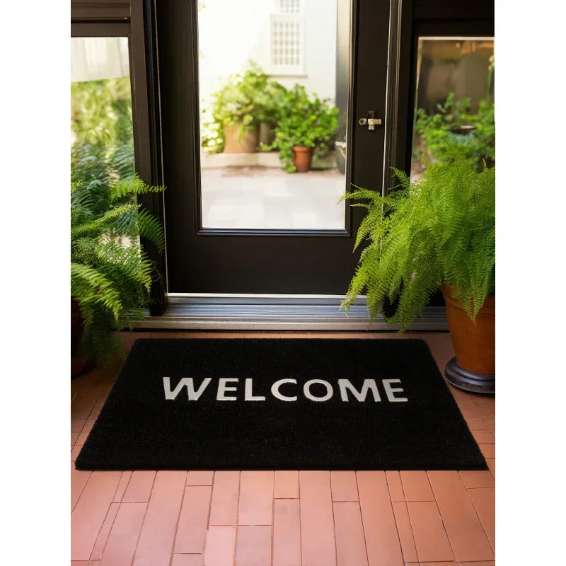 Black welcome doormat on terracotta tiled porch with potted ferns by entry door
