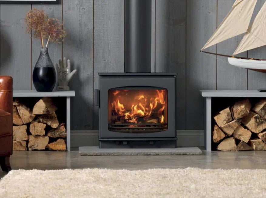 Black wood burning stove with fire, logs stacked on both sides, grey paneled wall backdrop