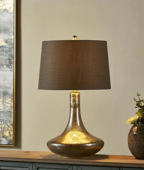 Gold glass table lamp with wide brown fabric shade on wooden sideboard