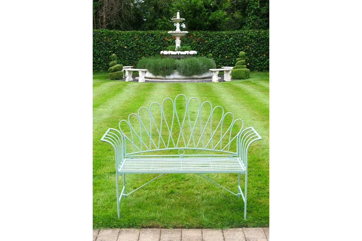 Metal garden bench with decorative arched backrest on striped lawn facing stone fountain