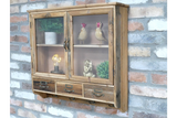 Wooden wall unit with glass doors, three small drawers, and metal handles, shown at slight left angle on brick wall.