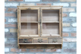 Wooden wall unit with two glass-front doors, three drawers, and four metal hooks, shown mounted on brick wall.