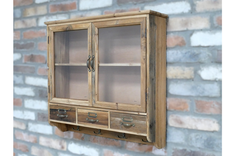 Wooden wall unit with glass double doors, three drawers, and four metal hooks, angled left view on brick wall.