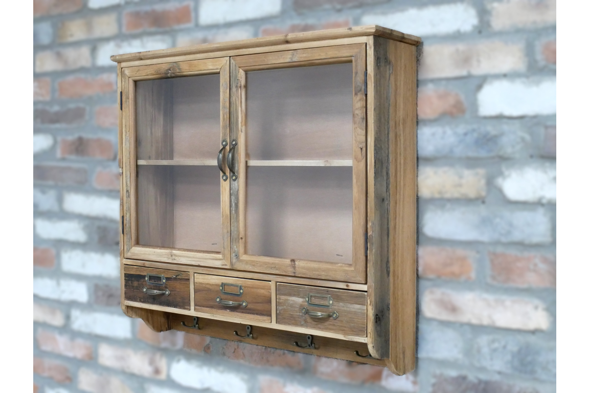 Wooden wall unit with glass double doors, three drawers, and four metal hooks, angled left view on brick wall.