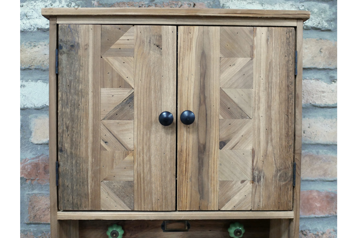 Wooden wall unit with two doors featuring geometric patterns, black knobs, and a lower open shelf against a brick wall.