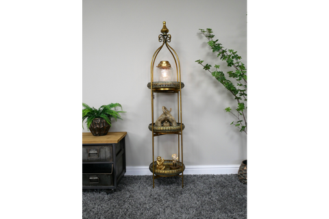 Tall narrow 3 tier metal shelves with gold finish, decorative trays, displayed in room with plants and grey carpet.