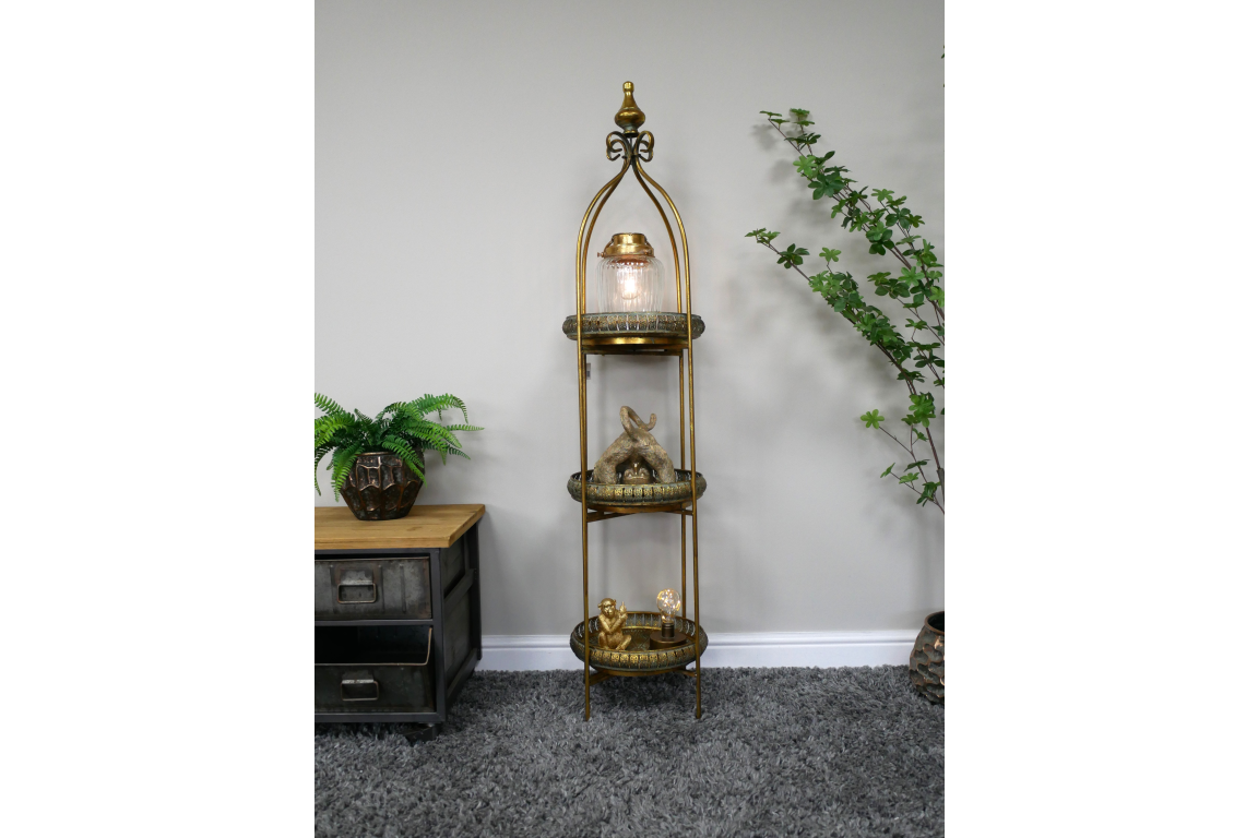 Tall narrow 3 tier metal shelves with gold finish, decorative trays, displayed in room with plants and grey carpet.