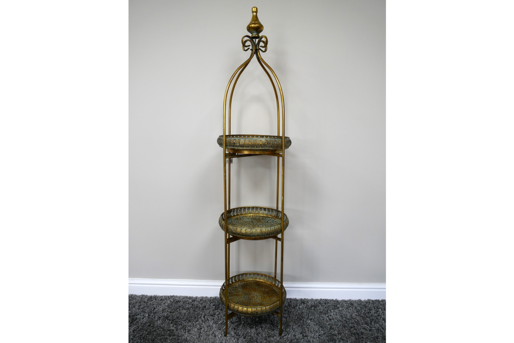 Tall 3 tier metal shelves with decorative round trays in gold and green, standing on a carpeted floor against a plain wall.