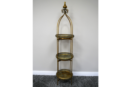 Tall 3 tier metal shelves with decorative round trays in gold and green, standing on a carpeted floor against a plain wall.