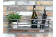 Grey metal wall-mounted shelf with railing, holding a small potted plant, wine bottle, and black figurine, against brick