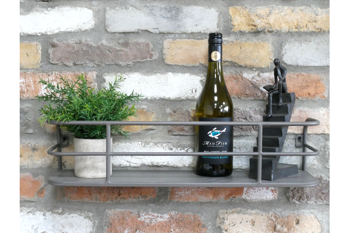 Grey metal wall-mounted shelf with railing, holding a small potted plant, wine bottle, and black figurine, against brick