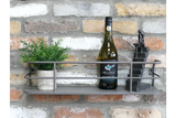 Grey metal wall-mounted shelf with railing, holding a small potted plant, wine bottle, and black figurine, against brick