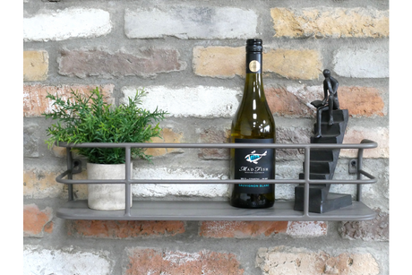 Grey metal wall-mounted shelf with railing, holding a small potted plant, wine bottle, and black figurine, against brick