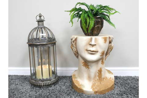 Beige and gold distressed resin head side table with a dark brown faceted planter holding a green fern plant, on grey carpet.