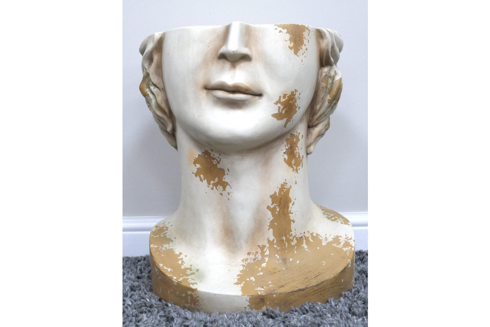 White and gold distressed ceramic head-shaped side table on gray carpet with white baseboard background