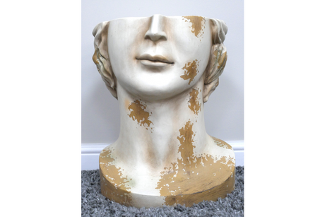 White and gold distressed ceramic head-shaped side table on gray carpet with white baseboard background