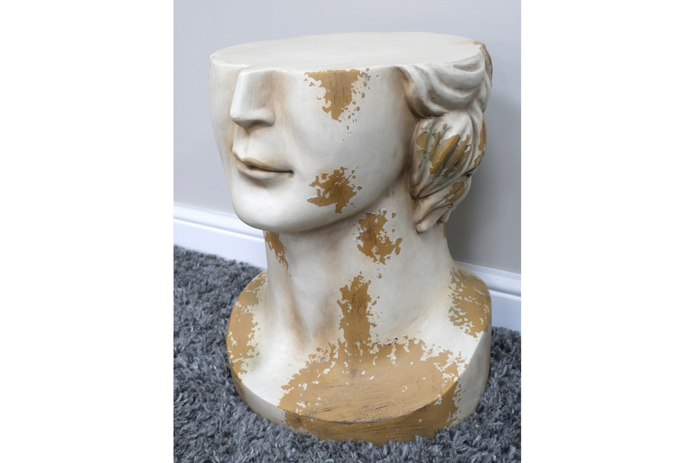 Side view of a white and gold distressed resin head-shaped side table on a grey carpet next to a wall.