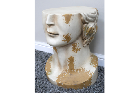 Side view of a white and gold distressed resin head-shaped side table on a grey carpet next to a wall.