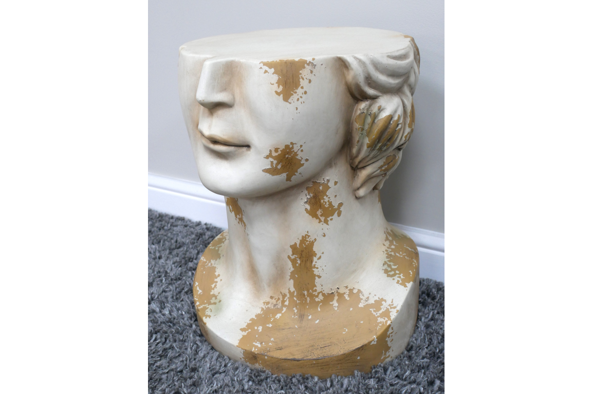 Side view of a white and gold distressed resin head-shaped side table on a grey carpet next to a wall.