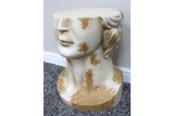 Side view of a white and gold distressed resin head-shaped side table on a grey carpet next to a wall.