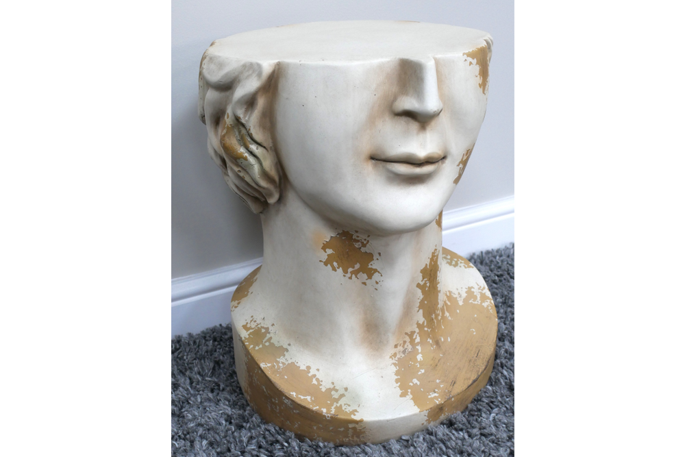 White and gold distressed ceramic head-shaped side table shown from front angle on gray carpet