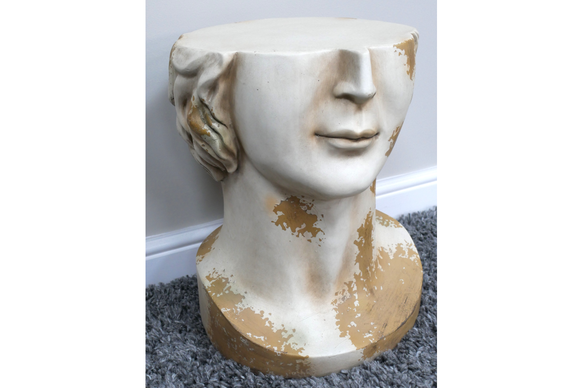 White and gold distressed ceramic head-shaped side table shown from front angle on gray carpet