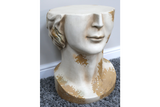 White and gold distressed ceramic head-shaped side table shown from front angle on gray carpet