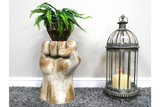 Side table shaped like a raised fist in distressed white and gold finish with a potted fern on top, next to a decorative