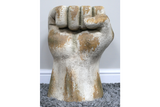 Side table shaped like a clenched fist, distressed white and beige finish, textured surface, standing on gray carpet.