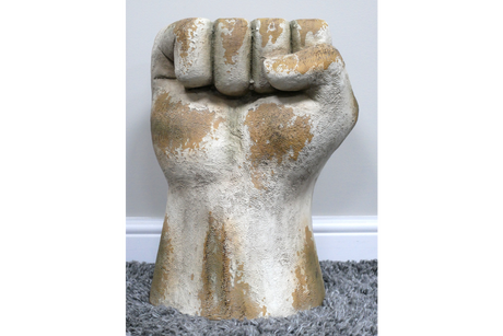 Side table shaped like a clenched fist, distressed white and beige finish, textured surface, standing on gray carpet.