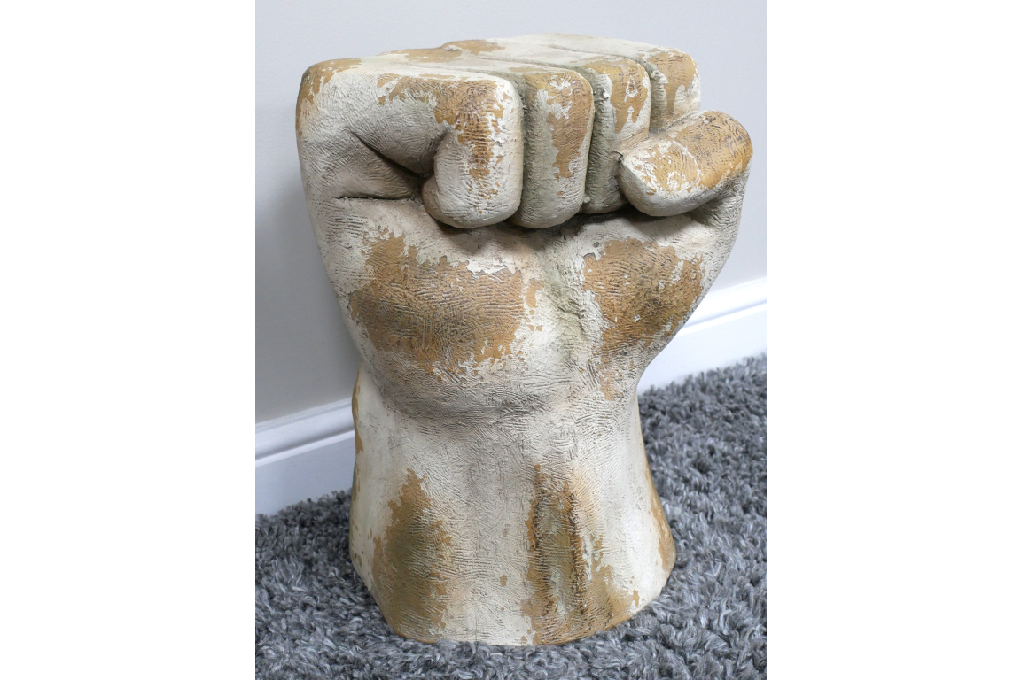 Fist-shaped side table in distressed white and beige finish, textured surface, shown on gray carpet with white baseboard