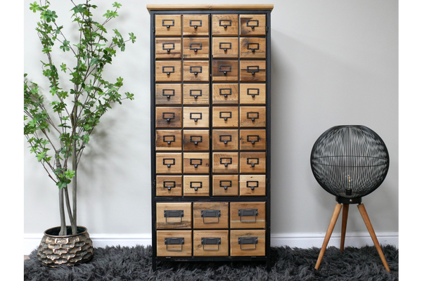 Tall wooden storage cabinet with multiple small drawers, black metal frame, standing on dark shaggy rug between plant and
